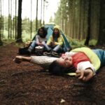 3 Menschen, die in einem Wald mit Schlafsack und Zelt übernachten.